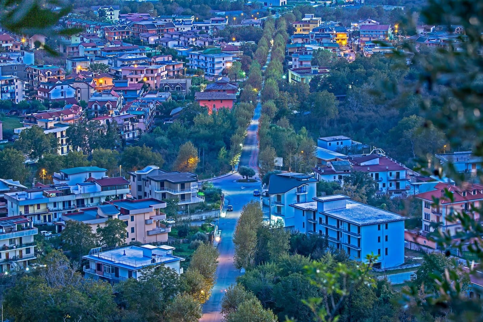 Un viale alberato che attraversa un paese al crepuscolo, visto dall’alto, con le luci accese e i toni blu della sera